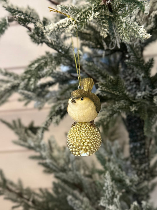 A gold-dusted bird ornament with a glittery body hangs gracefully from a branch of a snow-dusted artificial Christmas tree, adding festive charm to your holiday décor.