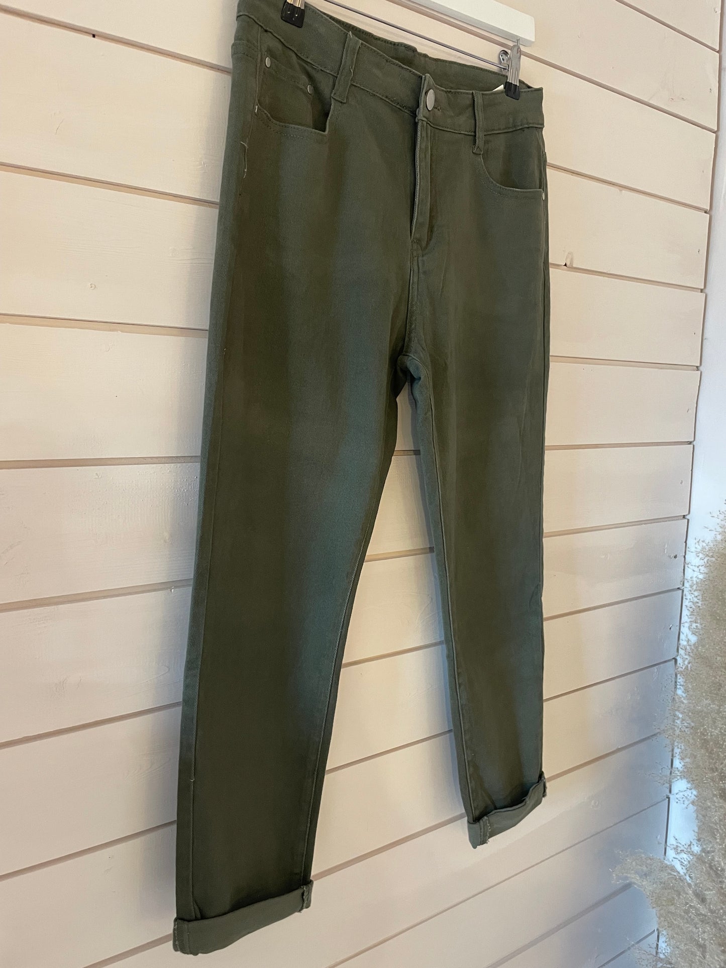 A pair of olive green cropped jeans with a shortened silhouette and turned-up cuffs hanging on a white wooden wall.