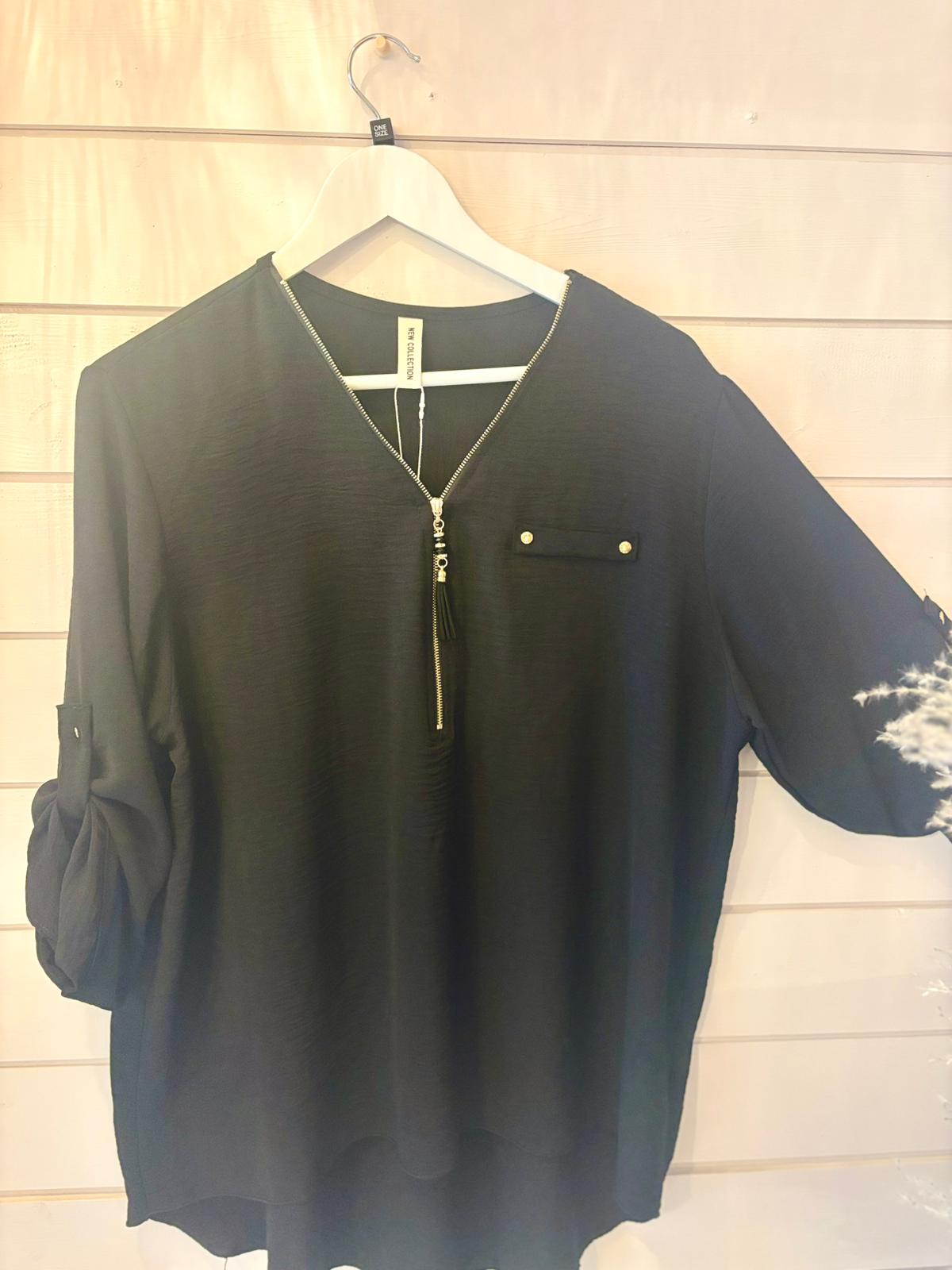 A black blouse featuring a zippered V-neck, chest pocket with button, and rolled-up sleeves, shown on a white hanger against a light wooden wall.