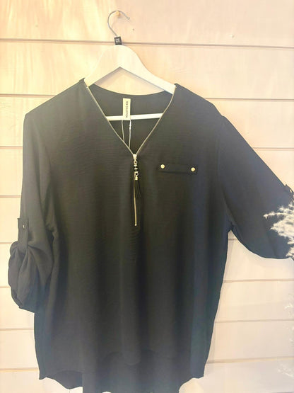 A black blouse featuring a zippered V-neck, chest pocket with button, and rolled-up sleeves, shown on a white hanger against a light wooden wall.