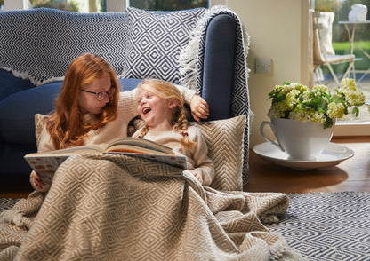 Wrapped in a cosy herringbone throw made from recycled plastic bottles, two young girls sit on the floor. One reads a book aloud, filling the air with laughter. Nearby, an eco-friendly cup-shaped planter blooms brightly.