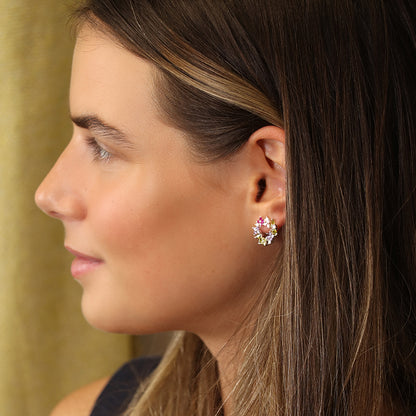 A woman with long brown hair is wearing pink and gold floral circle studs, facing left against a beige background.