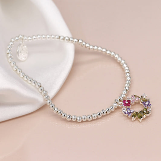 A necklace with silver beads and a flower-shaped pendant adorned with colourful gemstones lies on a reflective surface, reminiscent of an elegant floral crystal bracelet. A fabric is partially visible in the background.