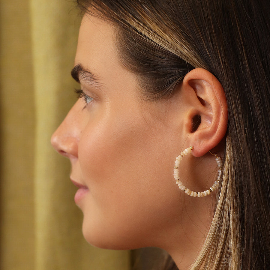 Side view of a person with long hair, adorned with hoops featuring seed pearls. The backdrop is a curtain.