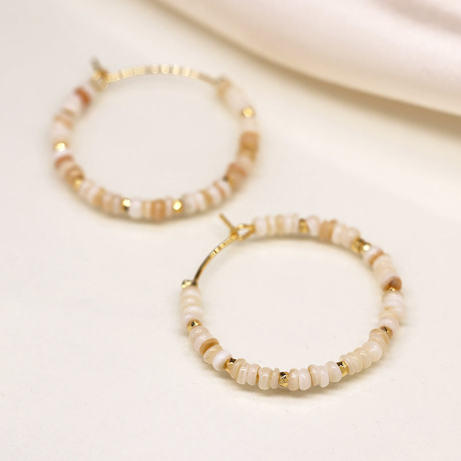 Close-up of two faux gold-plated hoop earrings adorned with small beige and white beads arranged in a circular pattern, resembling seed pearls, placed on a light-coloured surface.