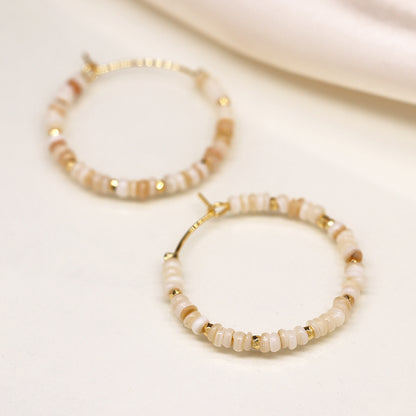 Close-up of two faux gold-plated hoop earrings adorned with small beige and white beads arranged in a circular pattern, resembling seed pearls, placed on a light-coloured surface.