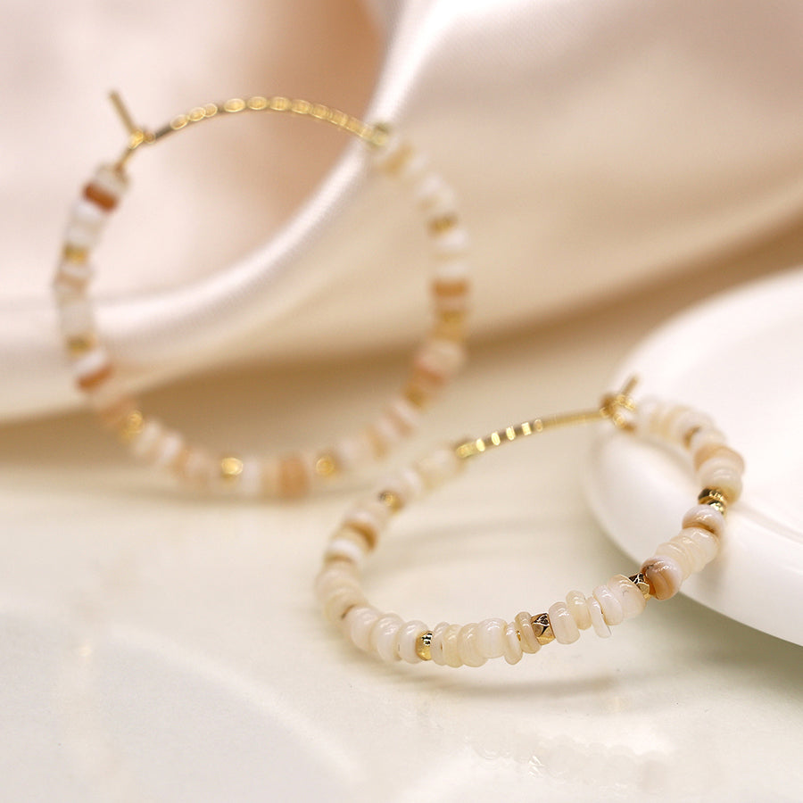 Two gold hoop earrings with beige and white beads elegantly rest on a cream surface, complemented by a silky fabric in the background. These faux gold-plated hoops add a touch of sophistication to any outfit.