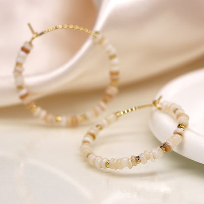 Two gold hoop earrings with beige and white beads elegantly rest on a cream surface, complemented by a silky fabric in the background. These faux gold-plated hoops add a touch of sophistication to any outfit.