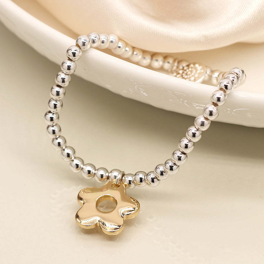 A silver-plated bracelet with a retro daisy charm rests elegantly on a white, scalloped dish against a faint satin cloth backdrop.