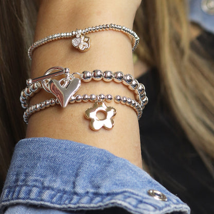 An individual adorned with multiple silver-plated bracelets featuring heart, flower, and retro daisy charms on their wrist. The individual is wearing a denim jacket.