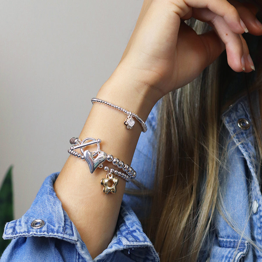A denim-clad person reveals their wrist adorned with silver-plated bracelets, one featuring a heart shape, another with a small pendant, and the third displaying a retro daisy charm.