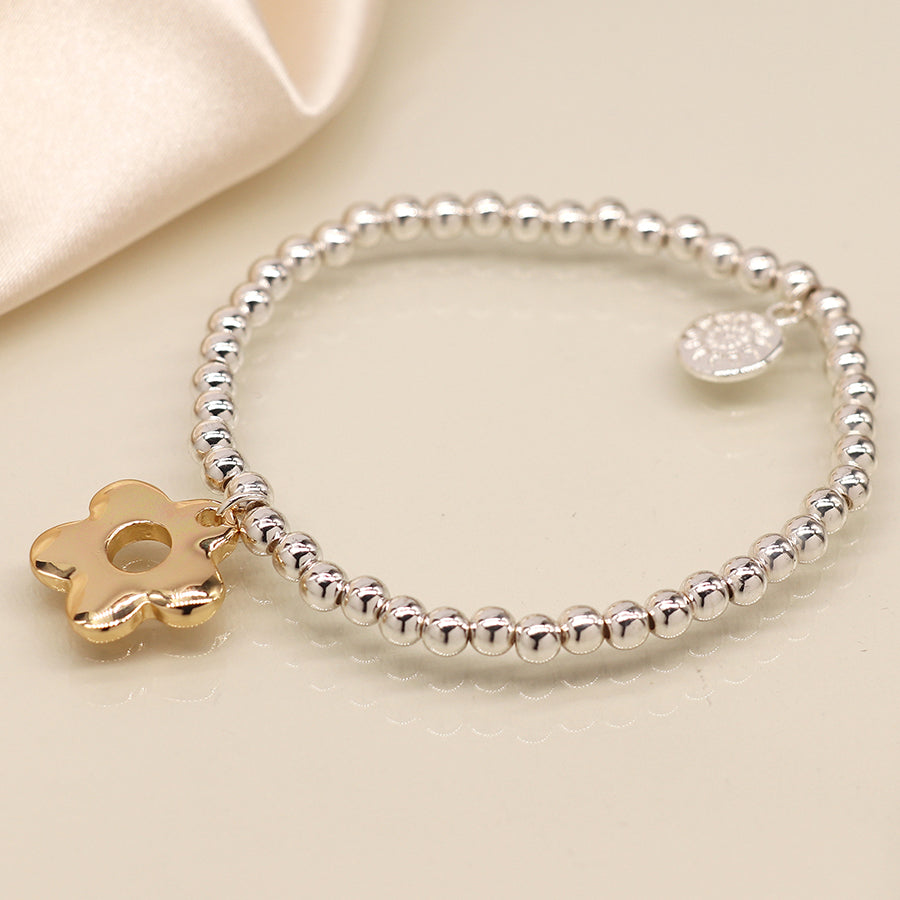 A silver-plated bracelet with a retro daisy charm and a small round tag lies on a beige surface.