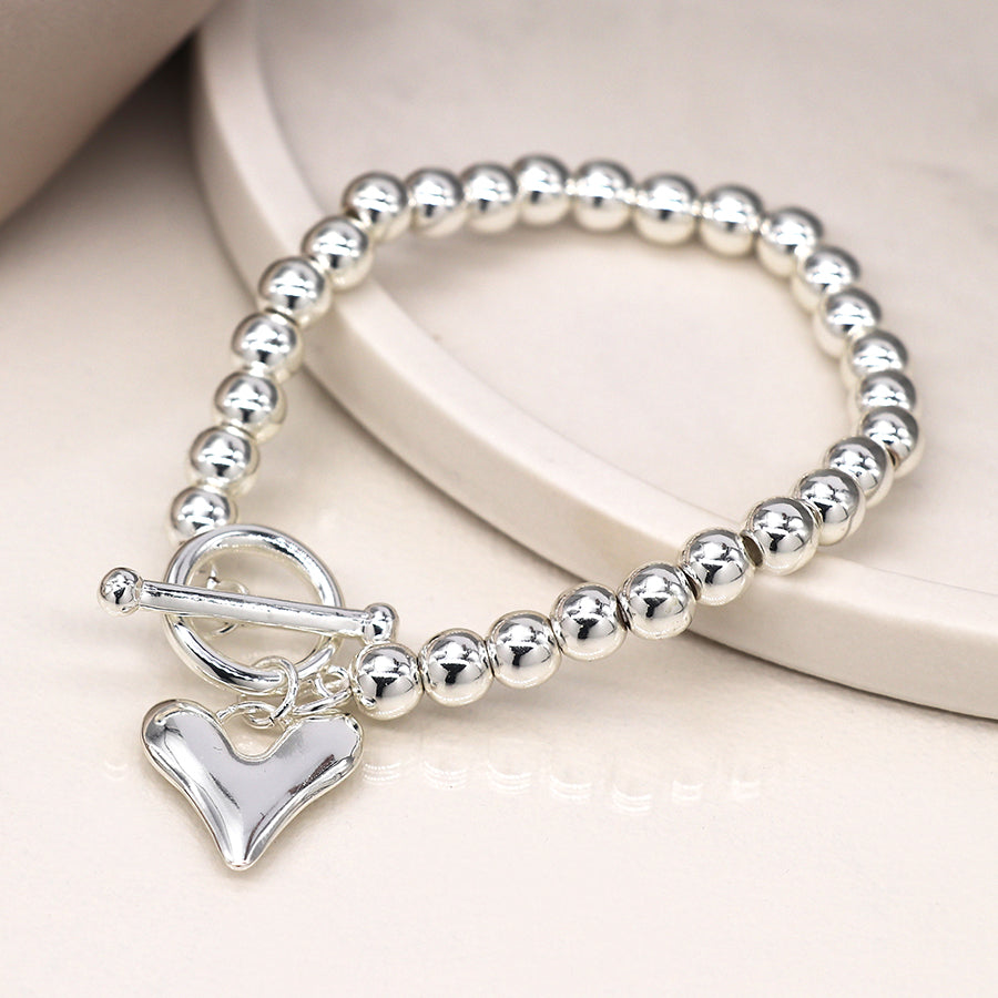 Silver-plated bracelet with round beads and a toggle clasp, featuring a heart-shaped charm, elegantly displayed on a light surface.