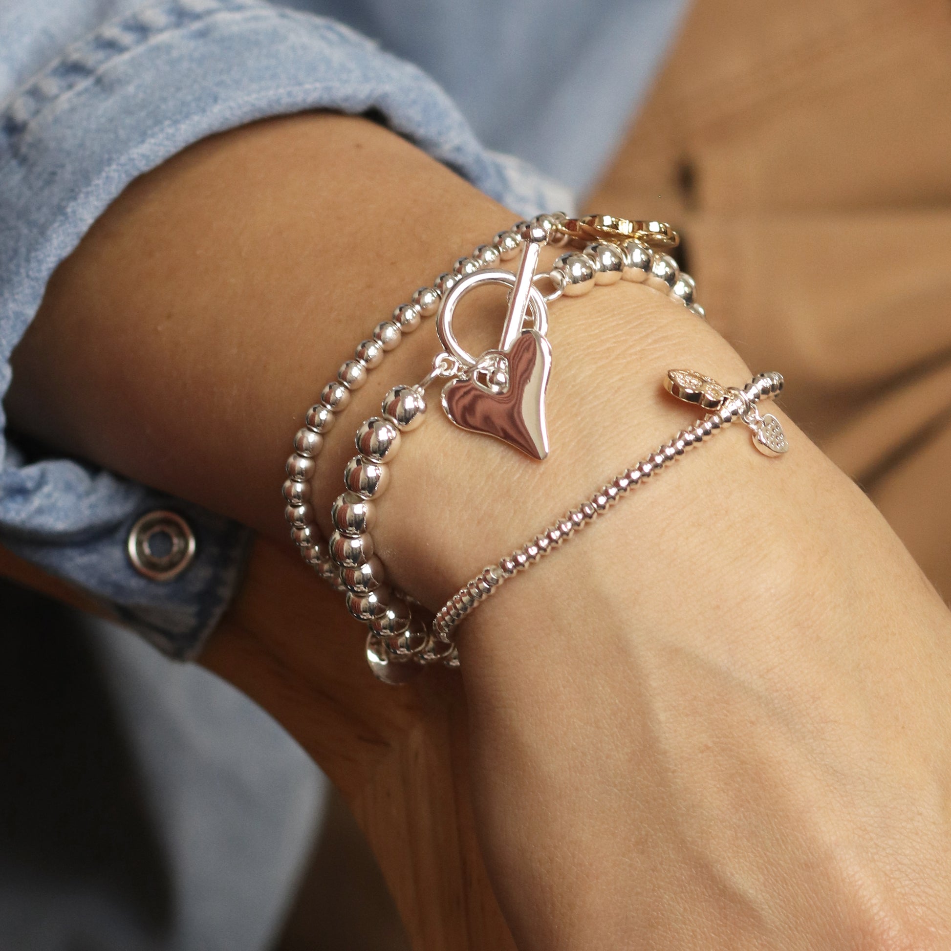A wrist adorned with a round bead stretch bracelet in silver and gold, featuring one with a heart charm and another with a small cross charm.