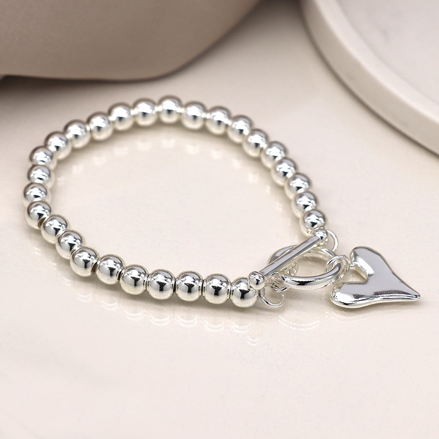 A silver-plated bracelet with a heart charm glistens on a reflective surface, showcasing round beads that elegantly complete the elasticated design.