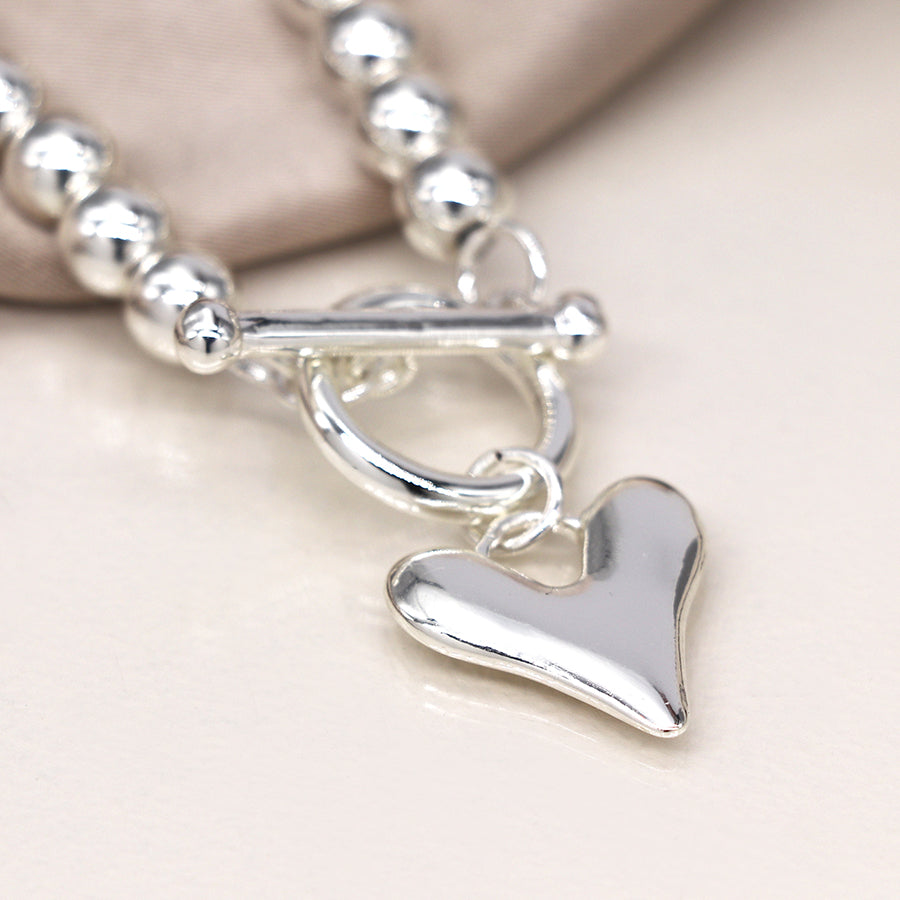 A silver necklace with round beads and a toggle fastening is adorned with a heart charm pendant, lying on a soft fabric background.