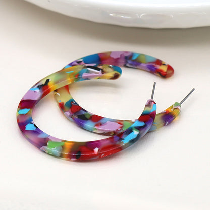 Colourful hoop earrings, a true statement piece, boast a confetti-like pattern with red, blue, green, and purple hues on a light surface.