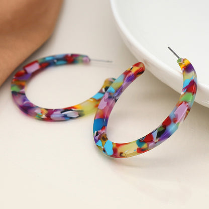 Two colourful acrylic statement piece earrings with a multicoloured mosaic pattern are placed on a white surface near a dish.