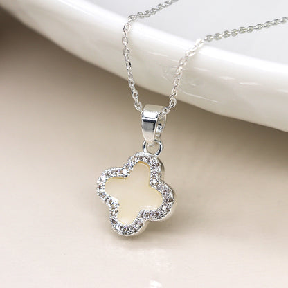 A silver-plated necklace featuring a crystal-edged, clover-shaped pendant with a cream-coloured centre, surrounded by small sparkly stones, displayed on a glossy white surface.