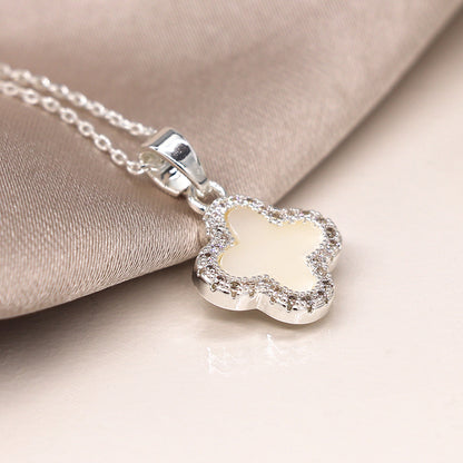 Silver-plated necklace with a clover-shaped pendant, featuring a white centre and a crystal-edged border. This exquisite jewellery piece rests elegantly on a beige fabric background.