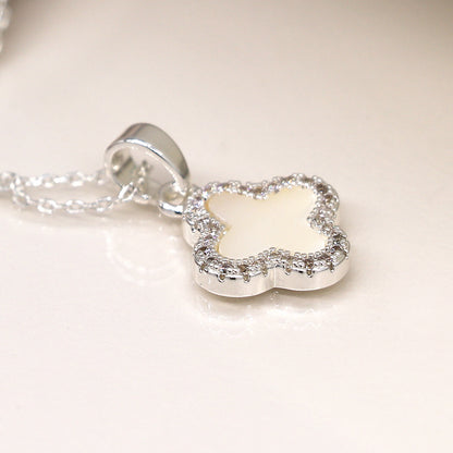 Silver-plated necklace with a flower-shaped pendant featuring a white centre and a border of small, sparkling stones, placed on a reflective surface. This piece exemplifies fine jewellery craftsmanship at its best.
