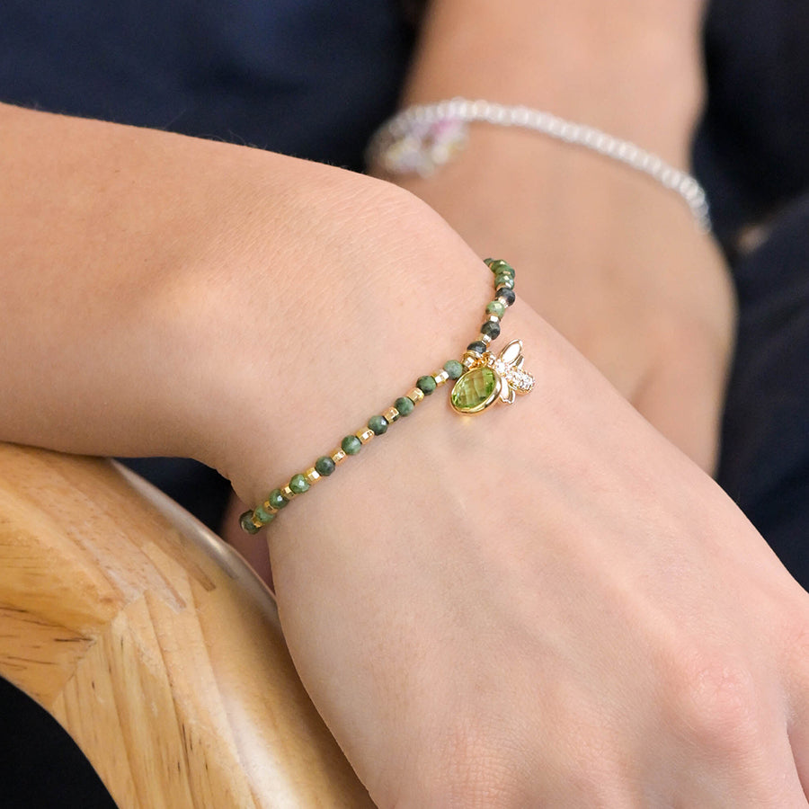 A person wearing a green and gold bracelet with a bee charm sits on a wooden chair, their wrist adorned by the fake gold-plated accessory.