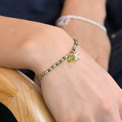 A person wearing a green and gold bracelet with a bee charm sits on a wooden chair, their wrist adorned by the fake gold-plated accessory.