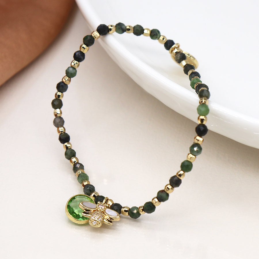 Green & Gold Bracelet adorned with a bee charm and faux gold-plated beads, featuring a small green pendant, elegantly rests on a white surface near a brown object.