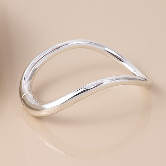 A smooth, curved silver-plated bangle with an open, wave-like design photographed on a beige surface, ideal for those who appreciate modern classic jewellery.