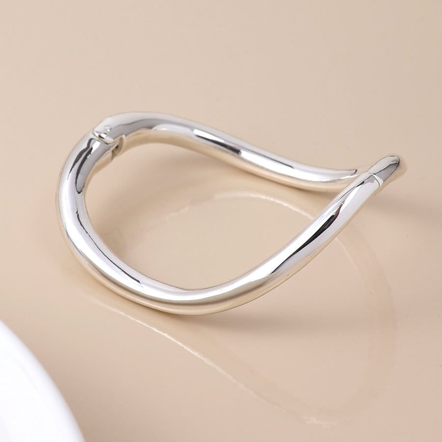 A polished, silver-plated bangle with an abstract form and curved edges lies on a beige surface—a modern classic piece of jewellery.