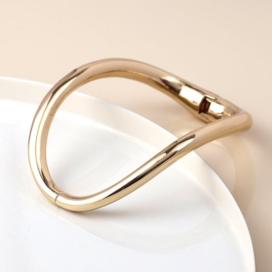 A gold hinged bangle with a wavy design lies partly on a white ceramic plate against a beige background—a timeless accessory, ideal for stacking.