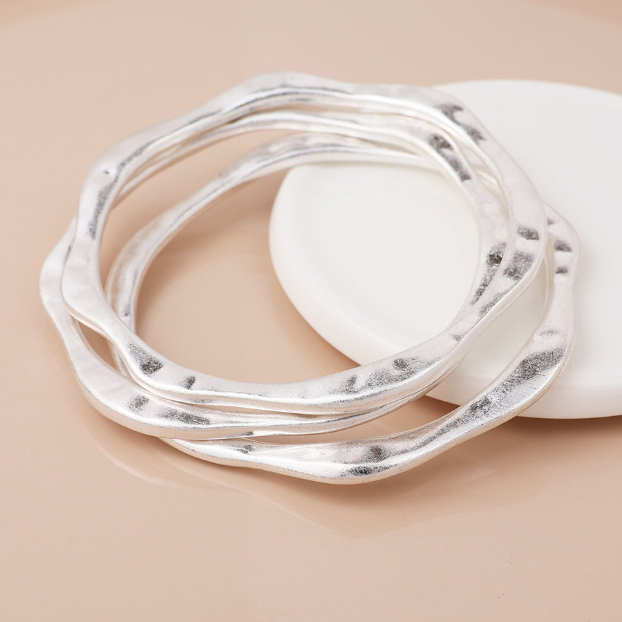 Three silver-plated bangles with a wavy-edged design are stacked on a round white dish set against a beige surface, creating an elegant layered appearance.
