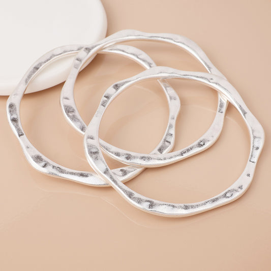 Three irregularly shaped, hammered silver-plated bangles are displayed on a light beige surface beside a white ceramic dish, ideal for achieving a stylish layered look.