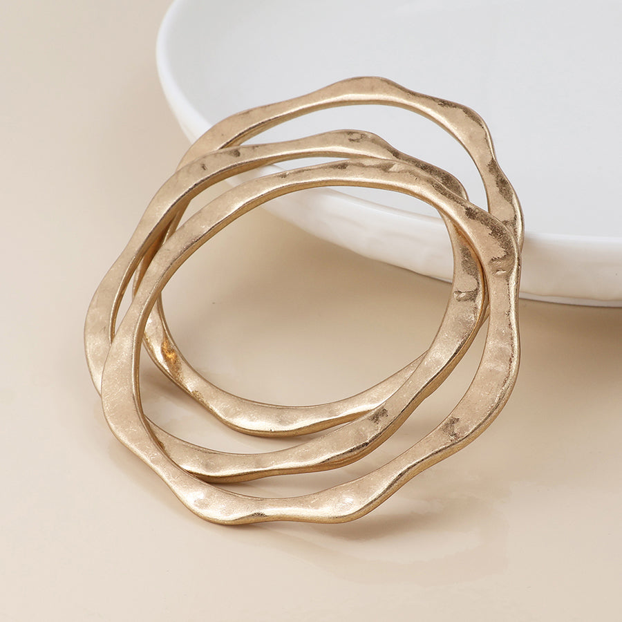 Three gold, wavy-edged bracelets are stacked together on a beige surface beside a white, round dish, bringing vintage charm jewellery to any ensemble.