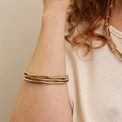 A person in a cream shirt showcases a gold chain necklace and three stackable bracelets, including the Golden Wave Bangle, adding a hint of vintage charm to their ensemble.