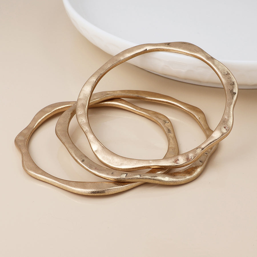 Three irregularly shaped gold bangles are stacked on a beige surface beside a white ceramic dish, creating an elegant gold bangle stack with a subtle vintage charm and jewellery appeal.