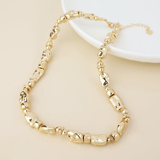 A gold beaded necklace featuring irregular, chunky beads and small round spacers, shown on a light surface beside a white dish—a versatile piece of jewellery suitable for any occasion.