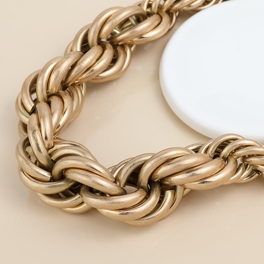 A close-up of a chunky, gold-toned chain necklace with large interlocking links is shown on a beige surface beside a round white object and a gold mixed bracelet.