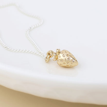 A gold, strawberry-shaped pendant adorned with small embedded gems hangs from a silver chain, forming a charming strawberry necklace displayed on a white surface.