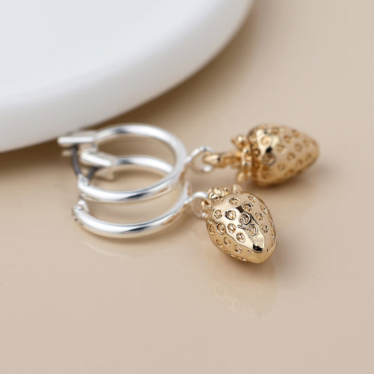 A pair of silver-plated hoops with gold strawberry-shaped charms rests on a beige surface next to a white object, bringing a playful touch of fruit-inspired jewellery to any outfit.