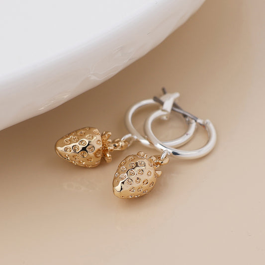 A pair of silver-plated hoops with gold strawberry-shaped charms offers a playful twist on fruit jewellery, resting elegantly on a light-coloured surface.