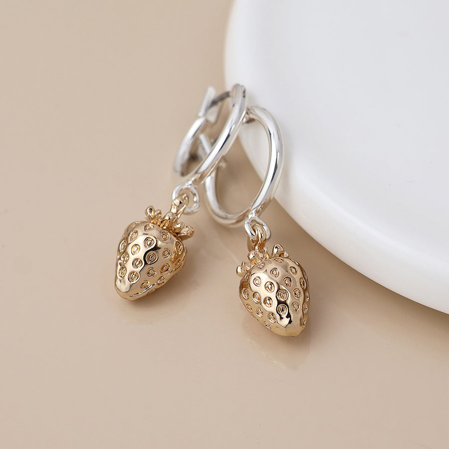 A pair of silver-plated hoops with gold, strawberry-shaped charms rests on a beige surface beside a white dish, combining elegance with a playful, fruity jewellery style.