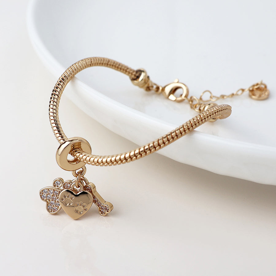 Ideal for animal lovers, this charm bracelet showcases golden crystals on a bear, a heart adorned with paw prints, and a bone, all presented gracefully on a white dish.