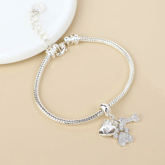 Silver paw charm bracelet with a snake chain design, featuring three dangling charms—a heart, a paw print and a bone—ideal jewellery for pet owners, displayed on a beige surface next to a white dish.