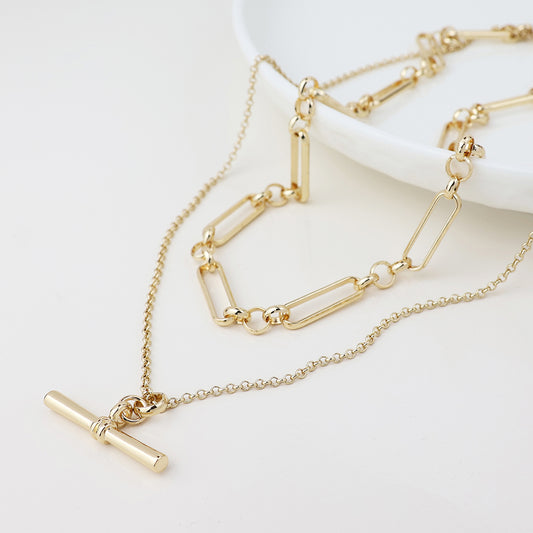 Two gold necklaces are shown on a white surface; one is a chic T-bar necklace, while the other has elongated and round links for a stylish, layered appearance.