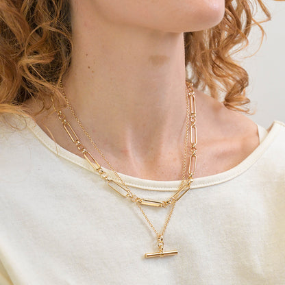 A person with curly hair wears a cream top and a gold layered necklace with a bar pendant.