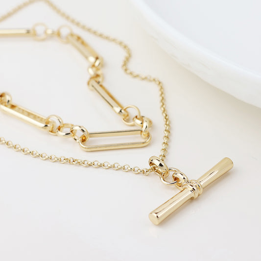 Gold layered necklace with rectangular links and a T-bar pendant, shown on a white surface close to the edge of a round dish.