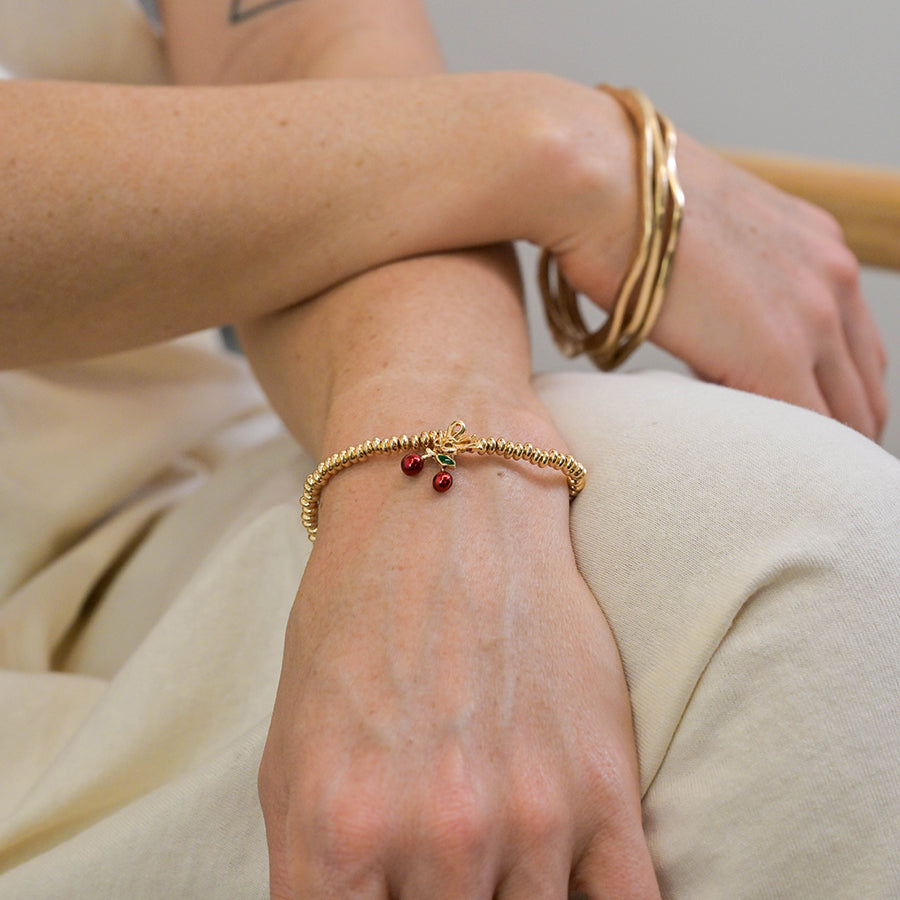 A person wearing a gold bracelet adorned with cherry charms and several gold bangles rests their arm across their lap.