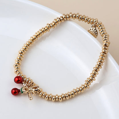 A gold beaded charm bracelet featuring two small red cherry charms and a golden bow-shaped charm, elegantly presented on a white plate.