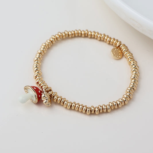 Gold beaded charm bracelet with a small red and white toadstool charm, shown on a white surface beside a round dish.